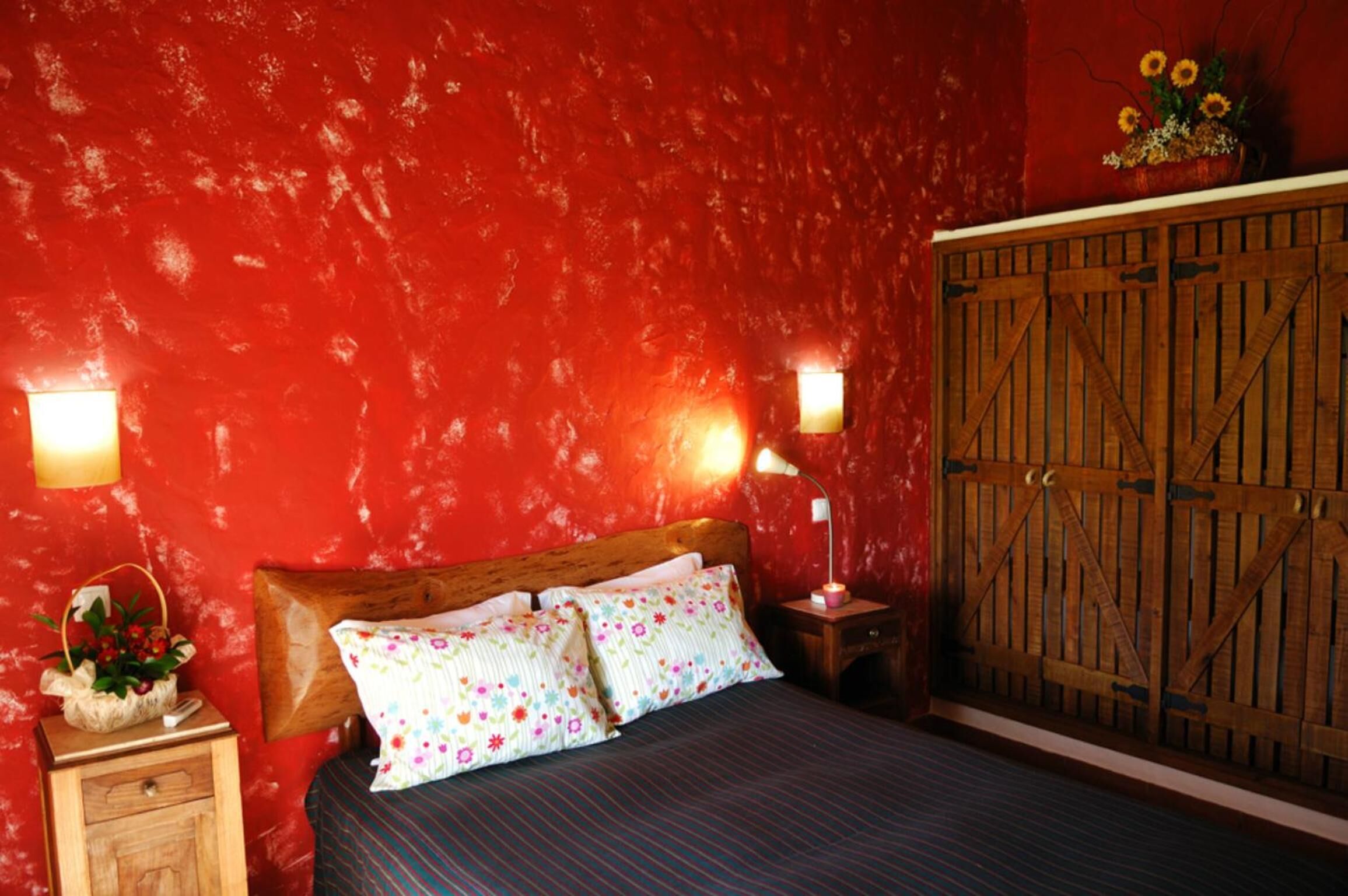 A bedroom with a red textured wall, a wooden bed with floral pillows, a nightstand with a lamp, and a wooden wardrobe.