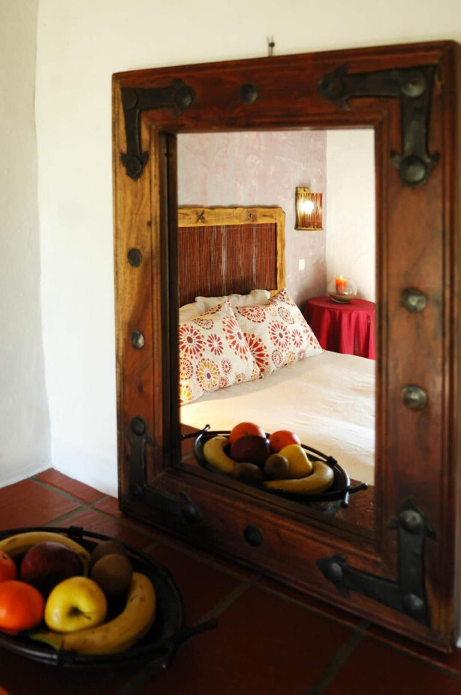 Een spiegel met houten lijst toont een bed met beddengoed met patroon en een fruitschaal op de vloer.