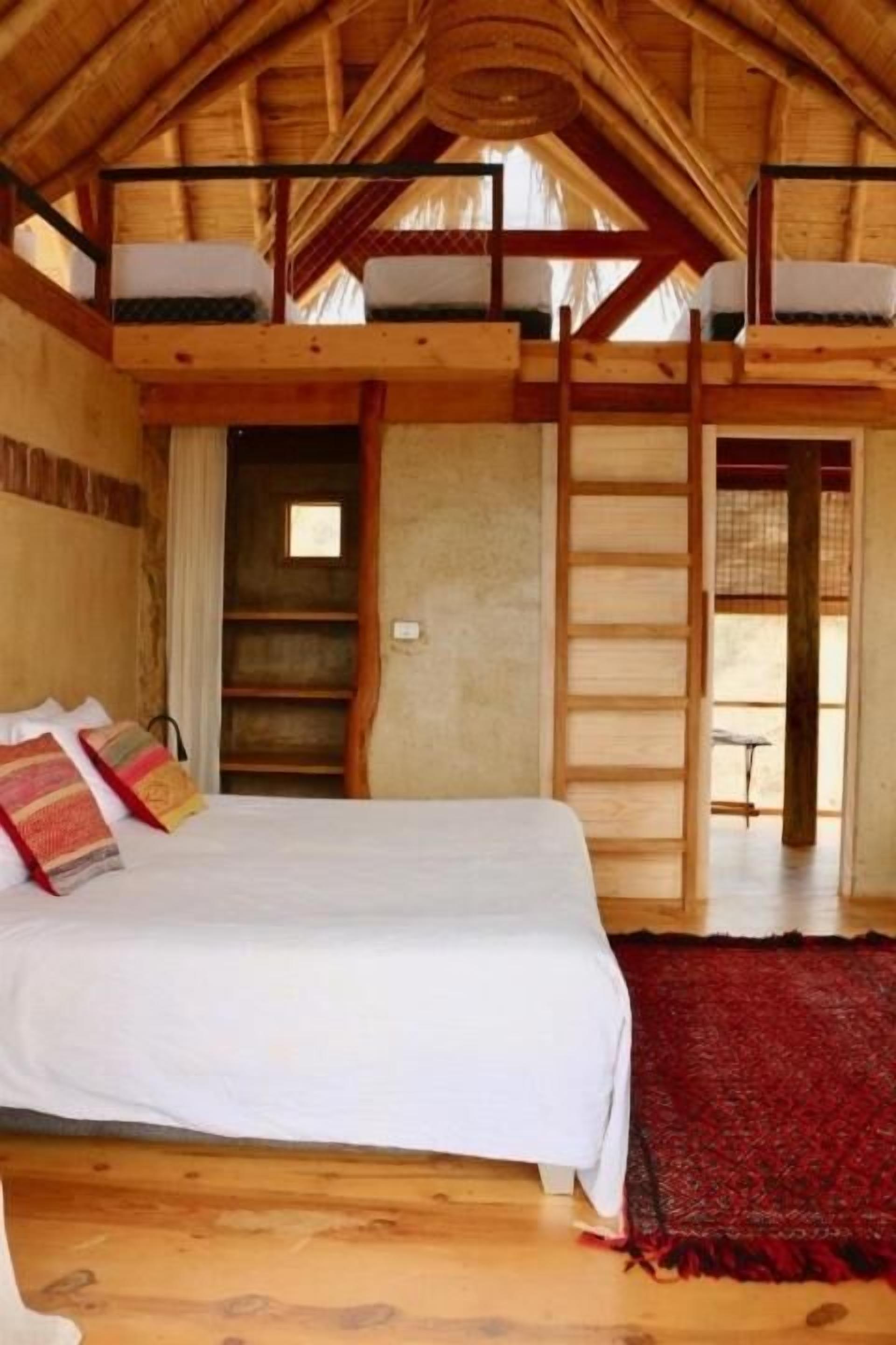 A room with a bunk bed, wooden beams, a ladder, a small window, and a red rug.