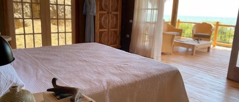 A bedroom with a large bed, a wooden floor, a view of the ocean, and a balcony with chairs.