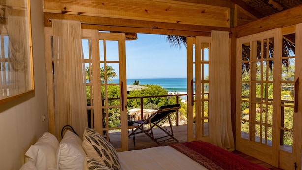 Une chambre avec un lit, un plaid rouge et une vue sur l’océan, visible à travers des portes en bois ouvertes.