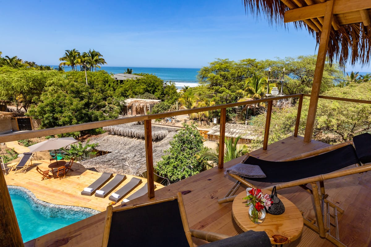 Terraza de madera con sillones de descanso y piscina, con vistas a una playa y al océano.