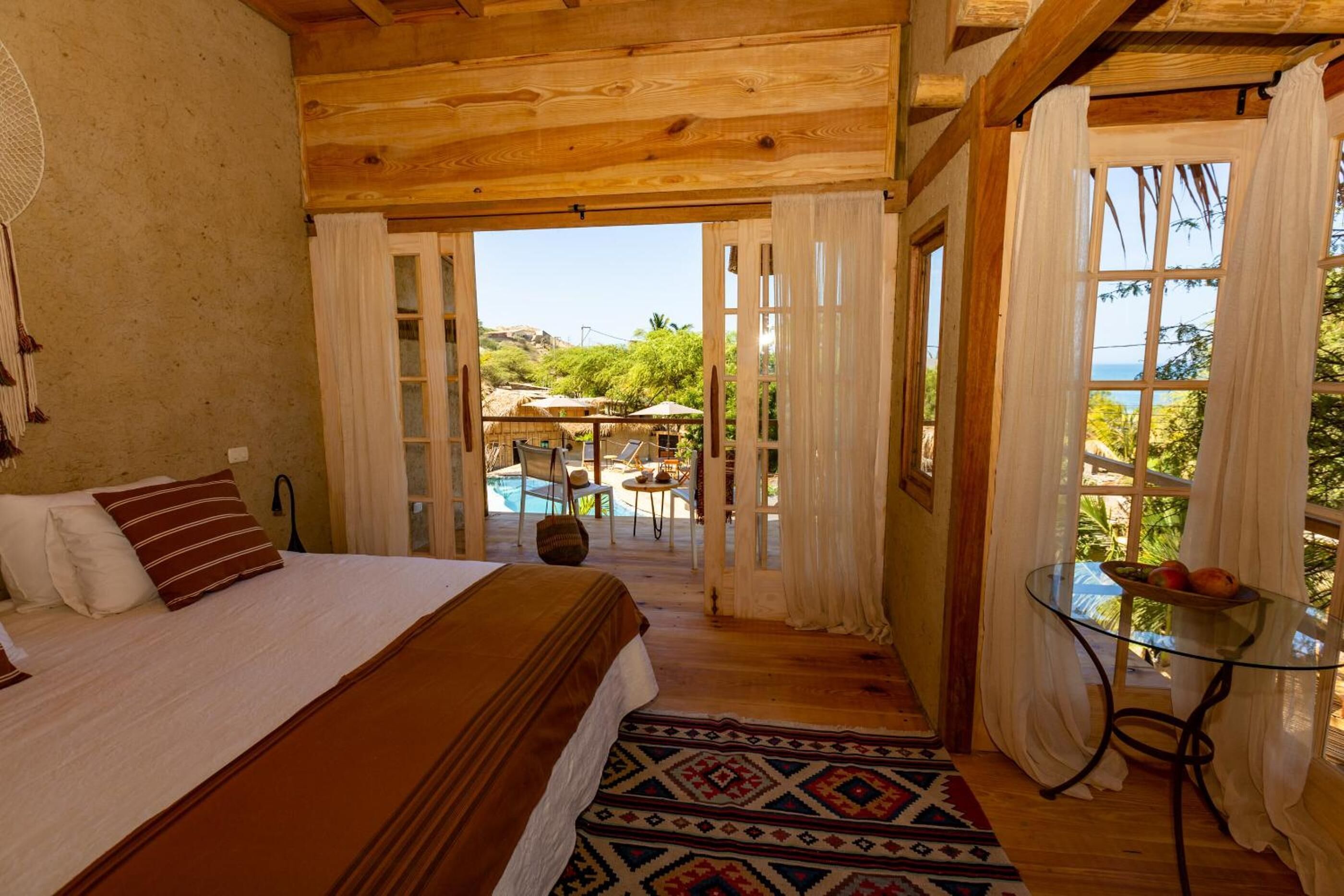 A wooden bedroom with a bed, a glass table with fruit, and a view of a pool and outdoor area.