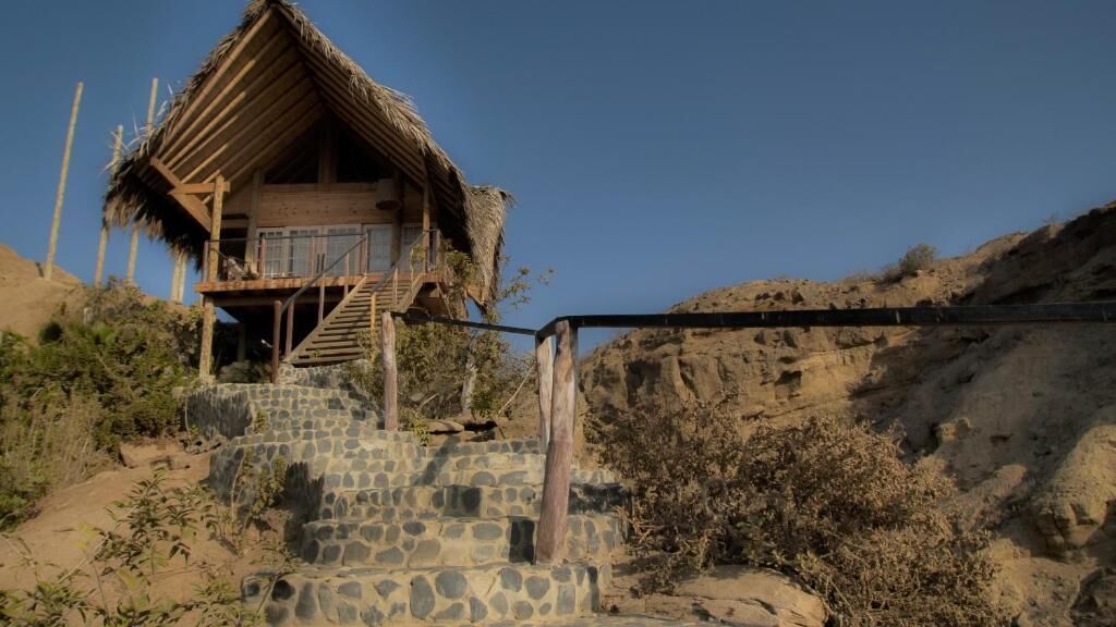 Un bâtiment rustique en bois, doté d’un toit de chaume, d’un escalier menant à un balcon et d’un mur de pierre au premier plan.