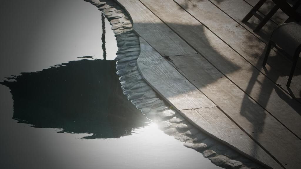 Une terrasse en bois bordée de pierres, une chaise et l’ombre d’une personne.