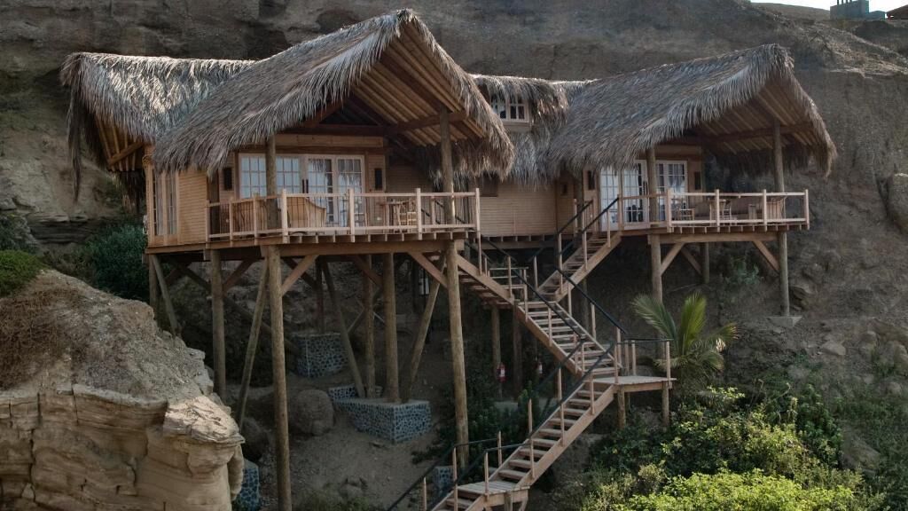 A wooden structure with a thatched roof, elevated on stilts, featuring a staircase leading to a balcony.