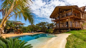 Une piscine entourée d’une terrasse en bois, de palmiers et d’un bâtiment à toit de chaume doté de balcons.