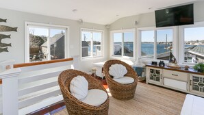 A spacious room with large windows, wicker chairs, a wooden bench, and a view of the sea. - Beautiful Hen Cove - Book Your 2026 Trip (Pocasset, Bourne)