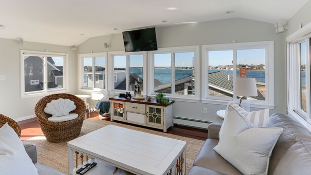 A spacious living room with a large window overlooking the sea, a white sofa, a wicker chair, and a white coffee table. - Beautiful Hen Cove - Book Your 2026 Trip (Pocasset, Bourne)