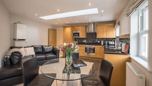 A modern kitchen with a dining area, featuring a glass table, dark leather sofa, and wooden cabinets. - Waterfront Apartments (Douglas)