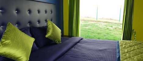 Una cama con cabecera capitonada, dos almohadas y vista a un área verde a través de ventanas amplias.
