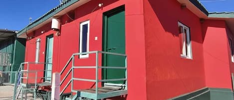 Ein rotes Gebäude mit grüner Tür und Fenstern, einer Metalltreppe und einer kleinen Außenfläche mit einem Stuhl.