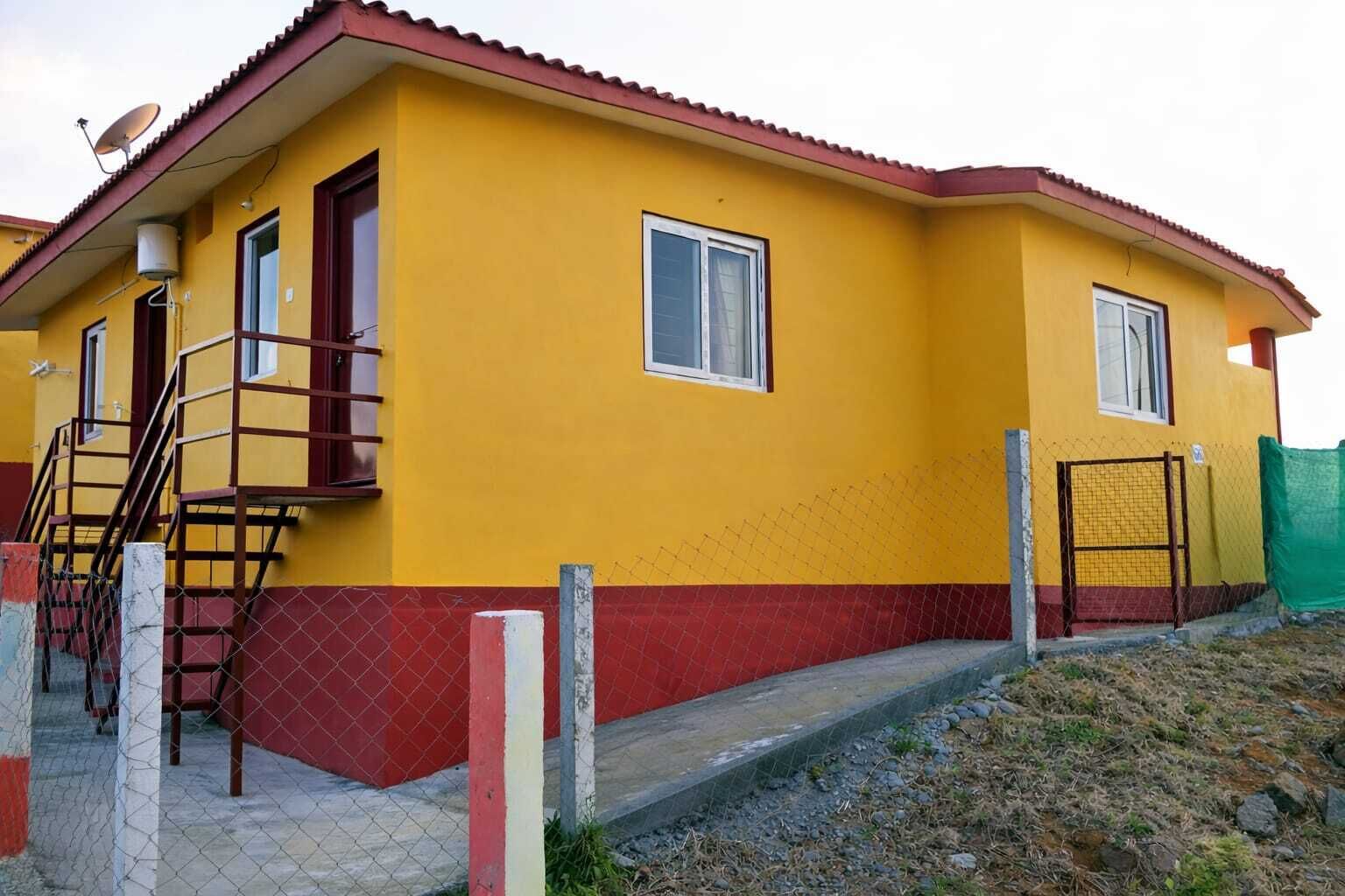 Un edificio amarillo con techo rojo, antena parabólica y entrada protegida por una reja.