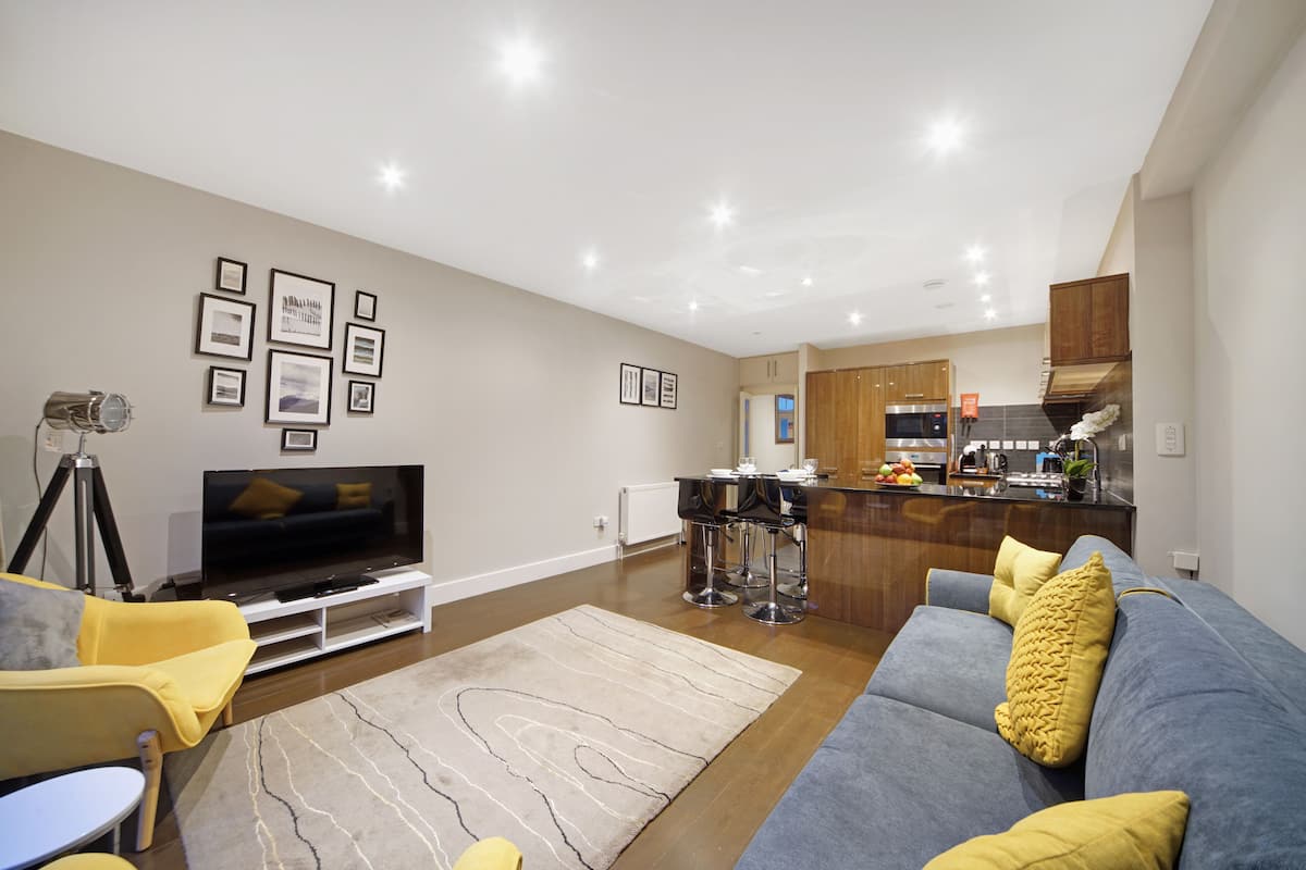 A modern living room with a flat-screen TV, a sofa, a coffee table, and a kitchen area in the background.
