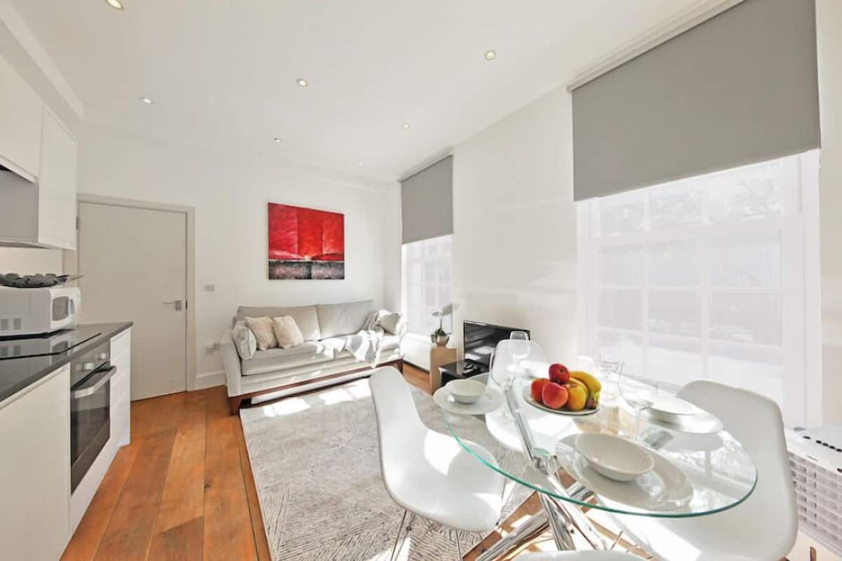 A modern kitchen with white cabinets, a black countertop, and a microwave. There is a dining area with a glass table, white chairs, and a fruit bowl. A sofa and a red abstract painting are visible in the background.