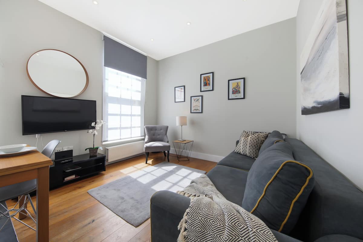 A modern living room with a grey sofa, a flat-screen TV, a dining table with chairs, and a large window providing natural light.