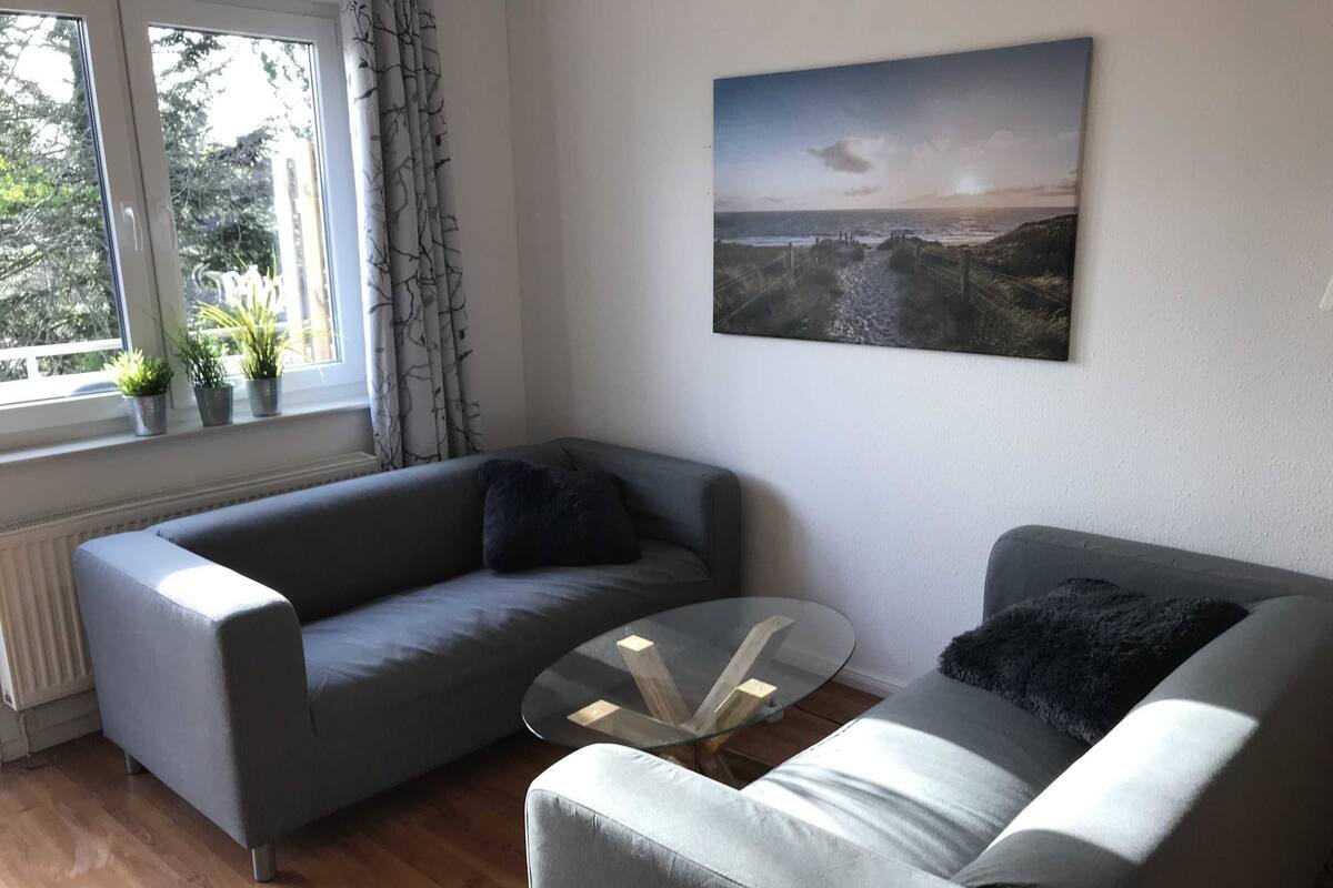 Una sala de estar moderna con dos sofás grises, una mesa de centro de vidrio y un cuadro de un paisaje costero.