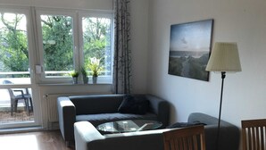 Una sala de estar con un sofá, una mesa de centro redonda de vidrio, una mesa de comedor de madera y sillas. Hay una ventana con cortinas, un radiador y una lámpara de pie.