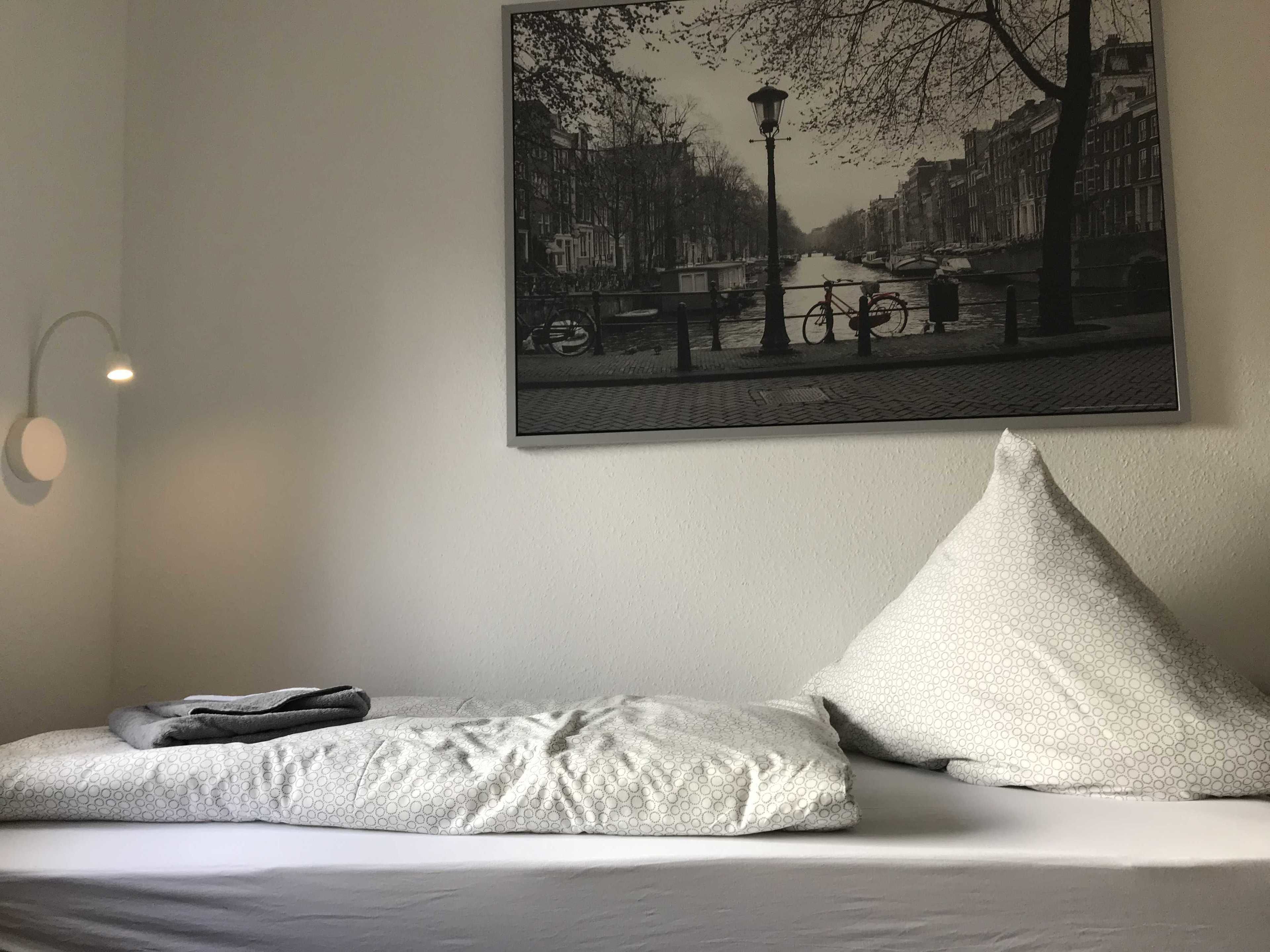 A bed with a pillow and a blanket, a wall-mounted picture of a canal, and a wall-mounted light fixture.
