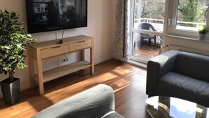 Una sala de estar con un televisor sobre un mueble de madera, un sofá gris, una planta en maceta y vistas a través de una ventana.