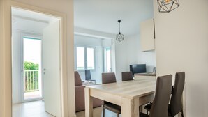 A dining area with a wooden table and chairs, a TV on a stand, and a balcony with a view. - Condominio Villa Mauro (Jesolo)