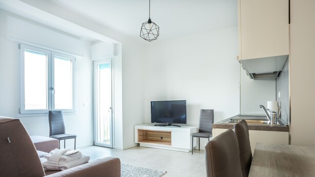 A modern living room with a flat-screen TV, a sofa, a dining table, and a kitchen area with a sink and cabinets. - Condominio Villa Mauro (Jesolo)
