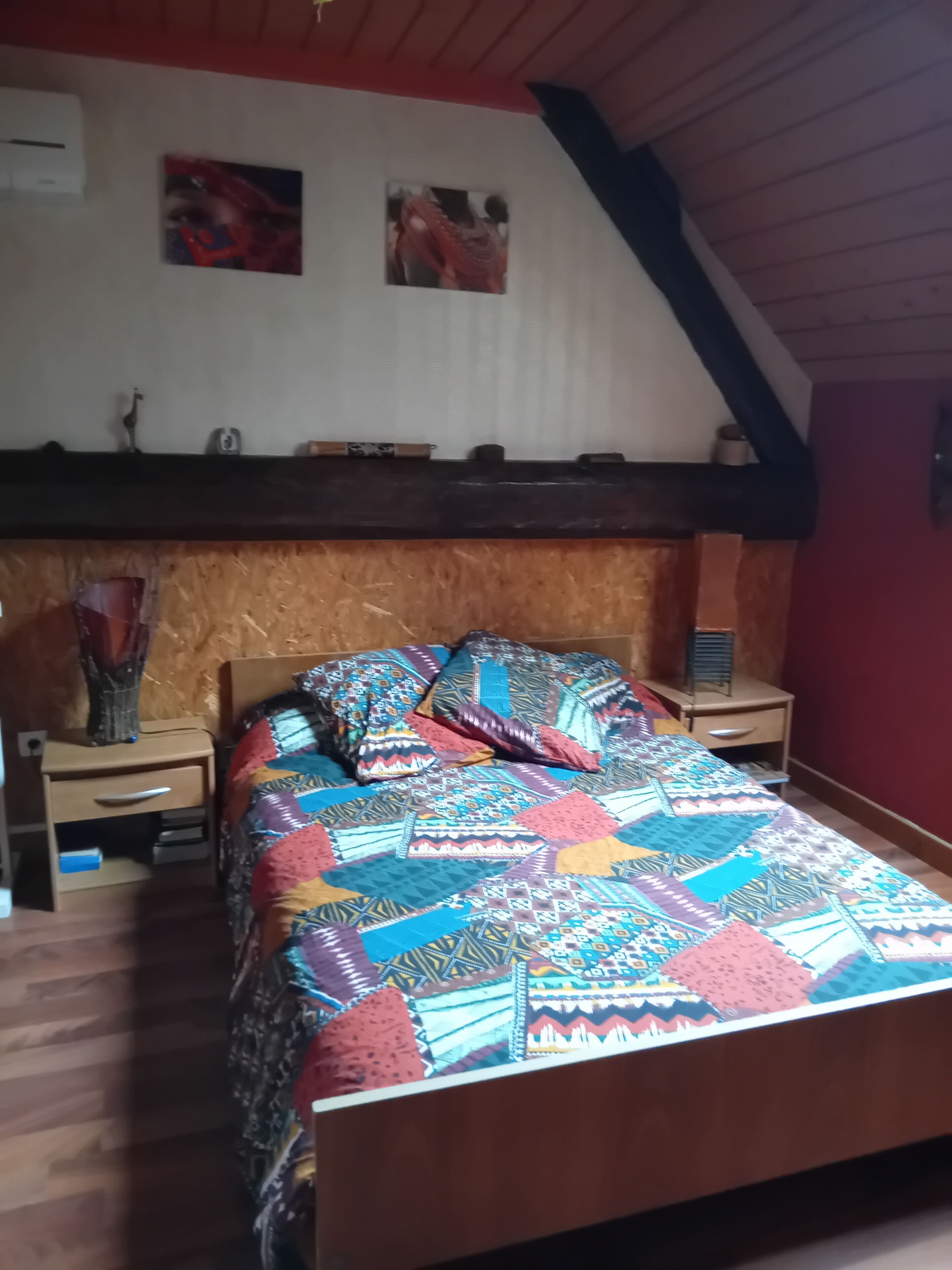 A bedroom with a wooden headboard, a colorful quilt, a nightstand with a lamp, and a small shelf with decorative items.