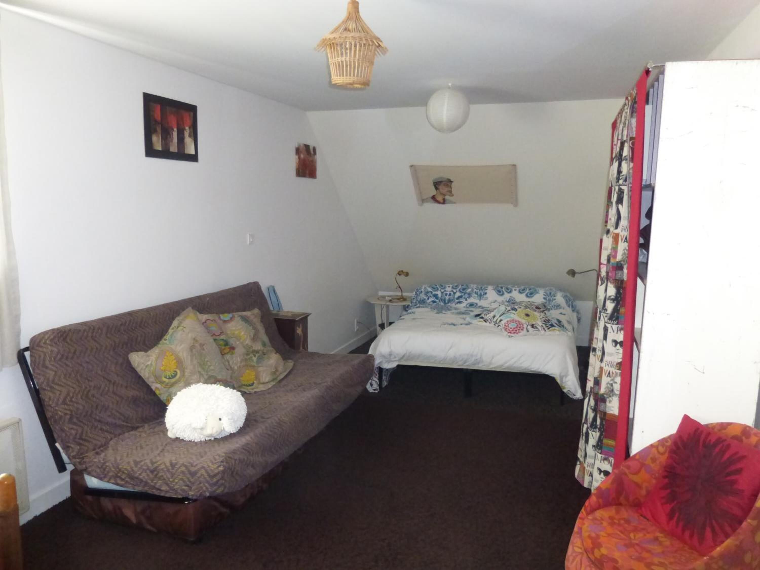 Um quarto pequeno e acolhedor com um sofá marrom, uma cama com colcha colorida, uma mesinha de cabeceira com abajur e um lustre.