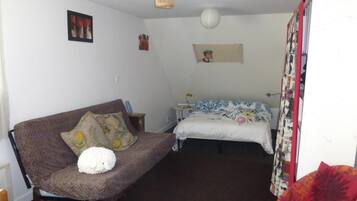 Um quarto pequeno e acolhedor com um sofá marrom, uma cama com colcha colorida, uma mesinha de cabeceira com abajur e um lustre.
