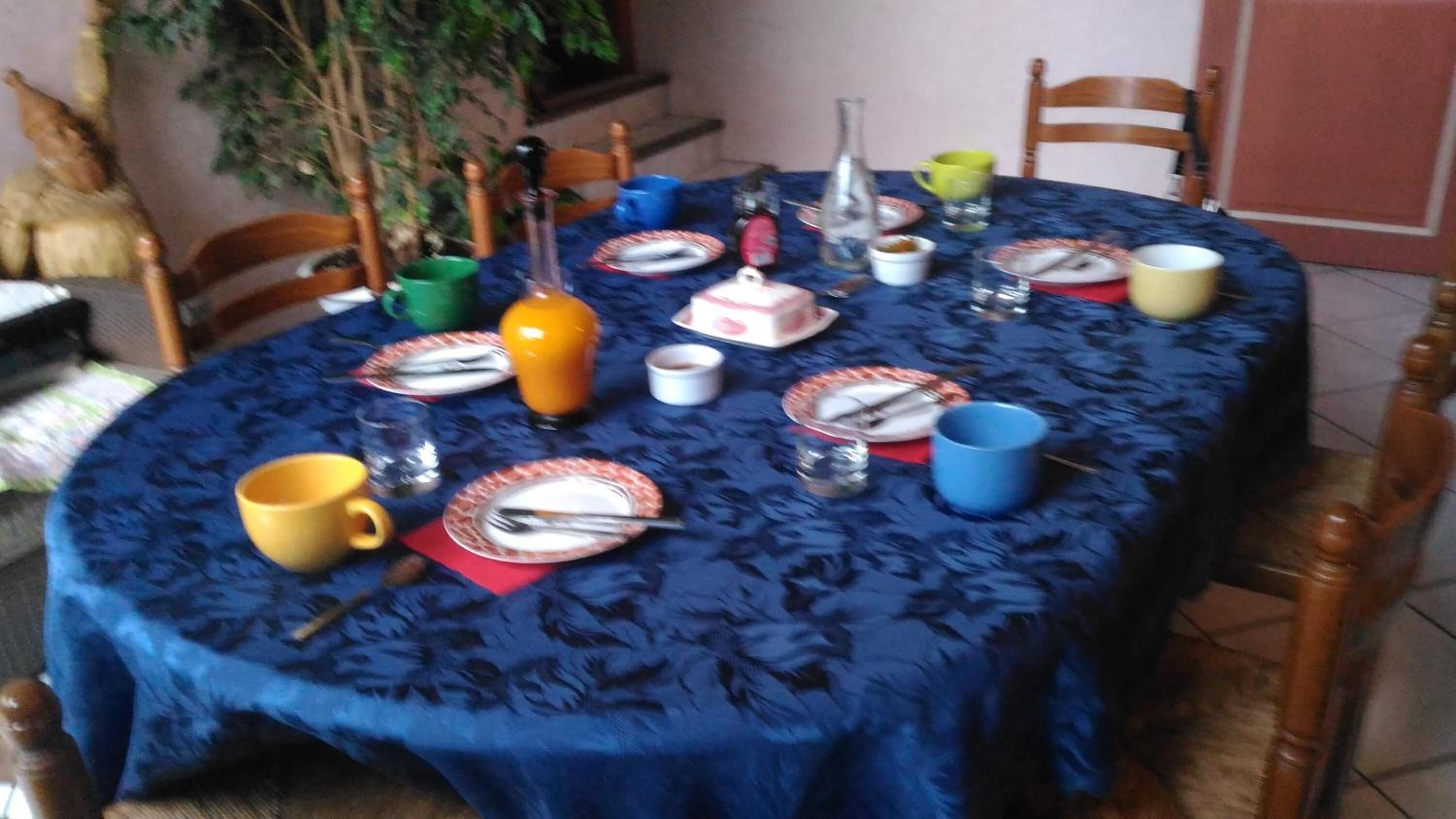 A round dining table covered with a blue tablecloth, surrounded by chairs, and set with plates, cups, and a bottle.