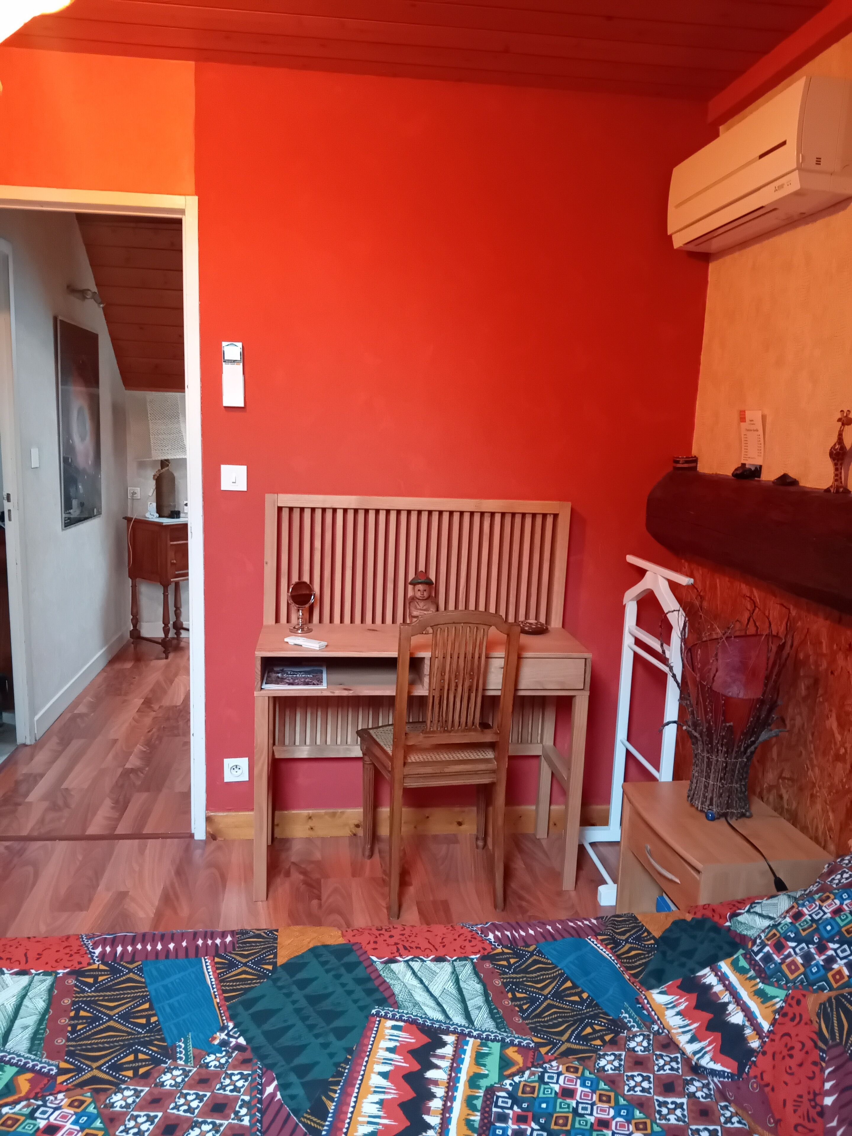 A room with a red wall, wooden desk, chair, and a bed with a colorful quilt.