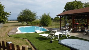 Un patio trasero con piscina, un muelle de madera y un área de patio techada con mobiliario de exterior.