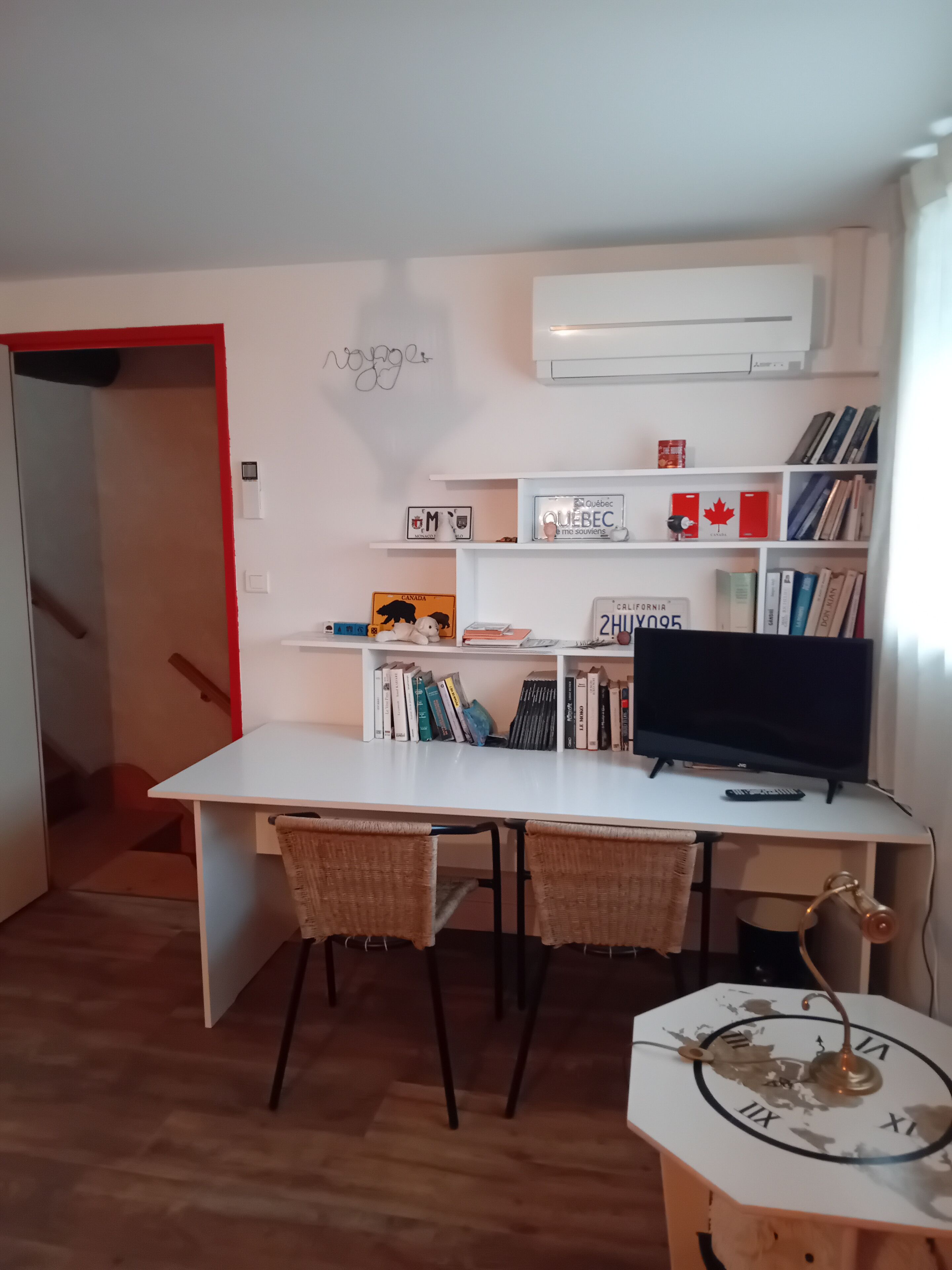 A study room with a desk, two wicker chairs, a bookshelf with books and decorative items, a television, and a wall-mounted clock.