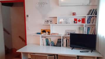 A study room with a desk, two wicker chairs, a bookshelf with books and decorative items, a television, and a wall-mounted clock.