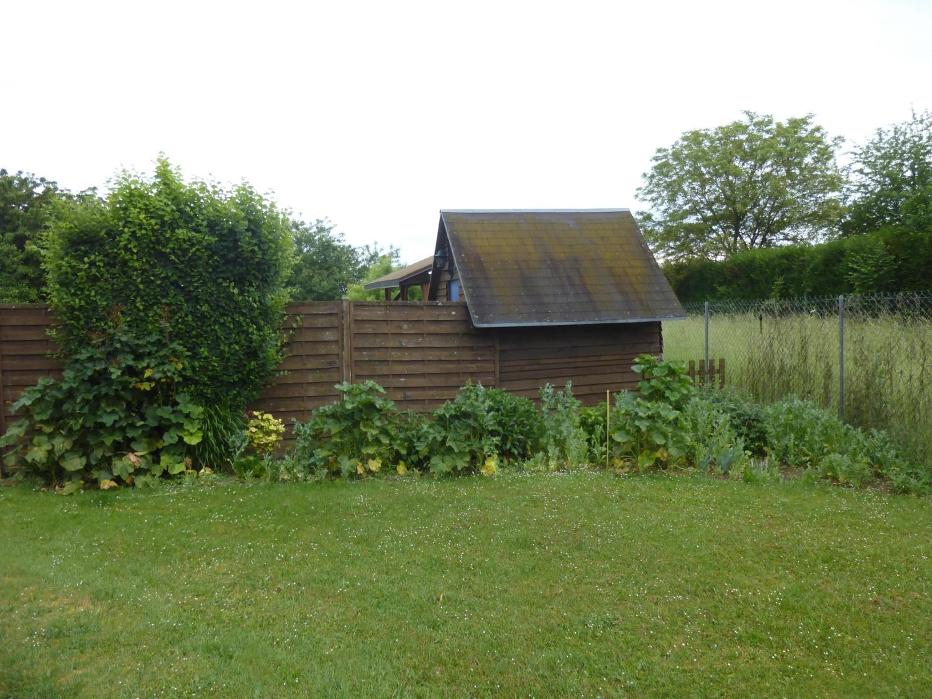 A rustic wooden structure with a slanted roof, surrounded by a lush garden and a wooden fence.