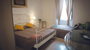 Un dormitorio con dos camas, una mesita de noche con lámpara, una pequeña mesa redonda con una planta y una mesa de centro de vidrio.