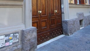 Una amplia puerta doble de madera con manija de metal, ubicada en una base de piedra, en un edificio de fachada amarilla y contraventanas marrones.
