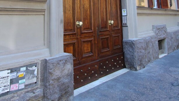 Una amplia puerta doble de madera con manija de metal, ubicada en una base de piedra, en un edificio de fachada amarilla y contraventanas marrones.