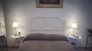 Una cama bien hecha con cabecera, dos mesitas de noche con lámparas, un cuadro en la pared y una planta pequeña a la derecha.