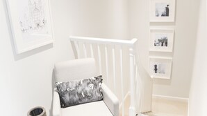 A white chair with a gray pillow, a white railing, and framed pictures on the wall. - The Apartment Bath (Bath)