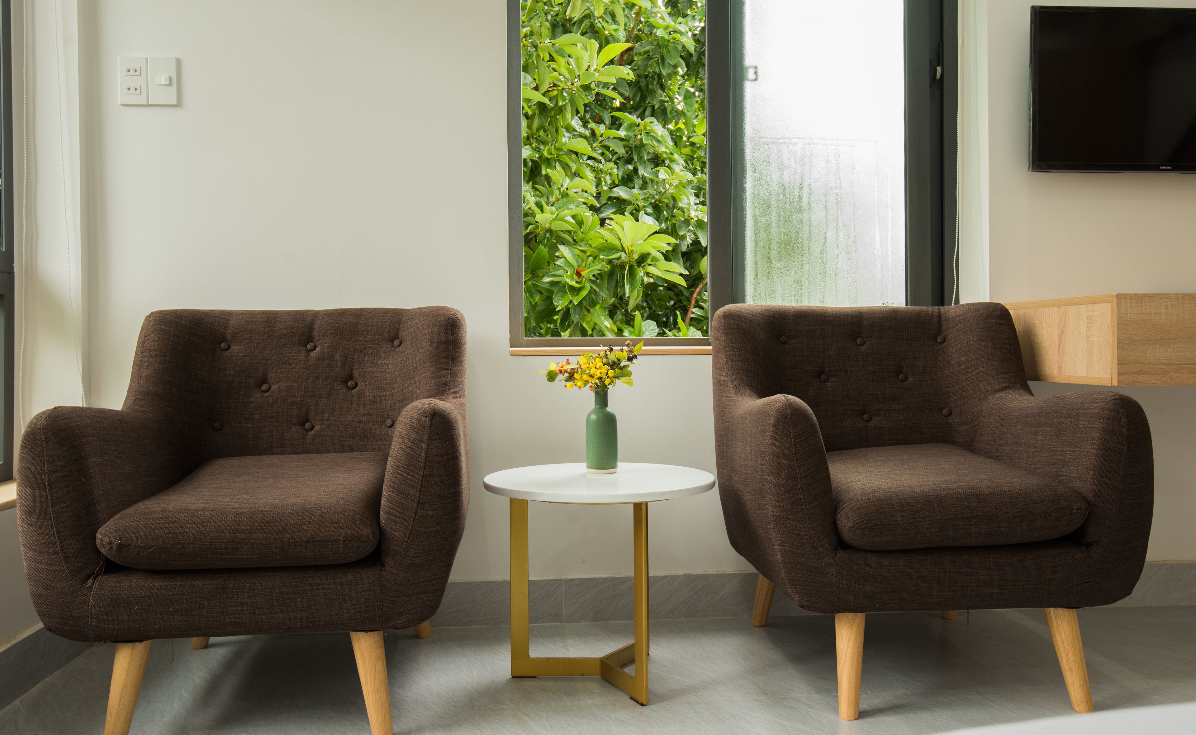 Two brown armchairs with wooden legs, a small white table with a green vase, and a window with a view of green foliage.