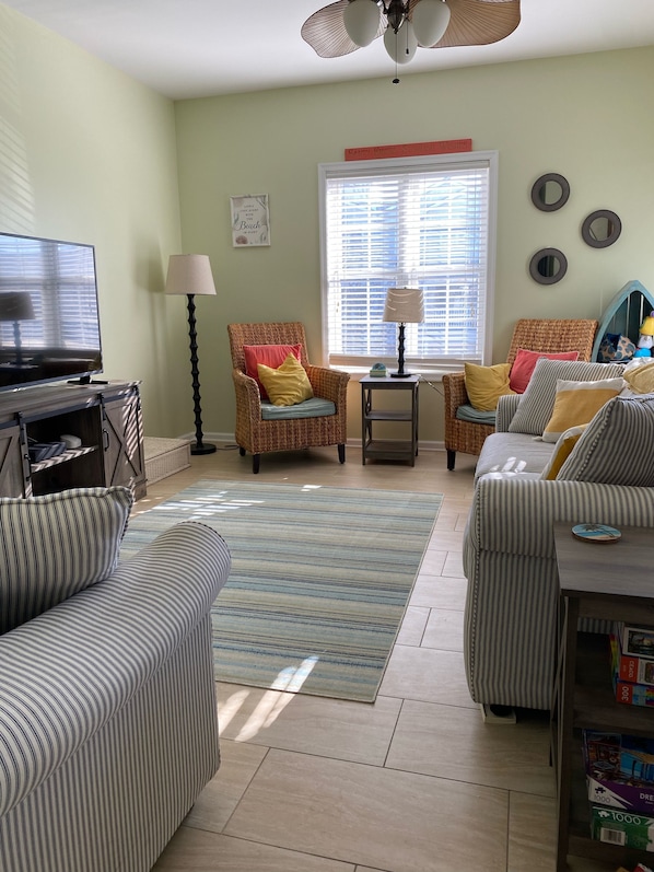 A living room with a television, wicker chairs, a striped sofa, a small table with a lamp, and a window with blinds.