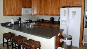 A kitchen with wooden cabinets, a microwave, a refrigerator, a sink, and a bar area with stools. - CLOSE TO ALL, a block from BEACH/BOARDS/ FAMILY FUN..Did we mention HEATED POOL! (Wildwood)