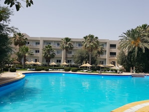 A swimming pool with lounge chairs and umbrellas, surrounded by palm trees and a building with balconies. - Azur Plaza (Hammamet)