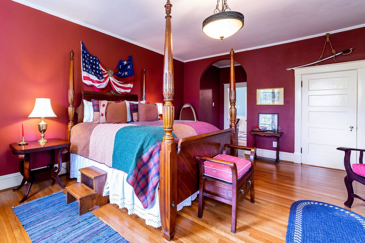 A bedroom with a four-poster bed, a wooden chair, a small table with a lamp, and a blue rug.