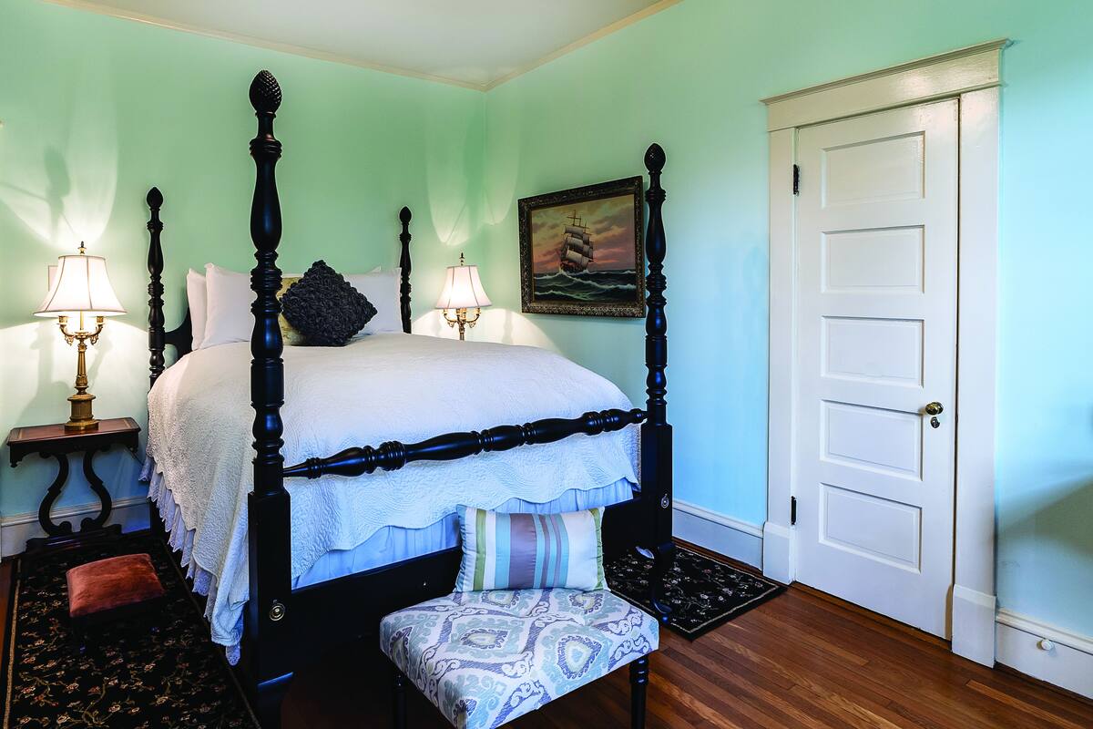 A four-poster bed with a white bedspread, a bench with a patterned cushion, two bedside lamps, and a framed picture of a ship on the wall.