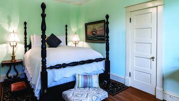 A four-poster bed with a white bedspread, a bench with a patterned cushion, two bedside lamps, and a framed picture of a ship on the wall.