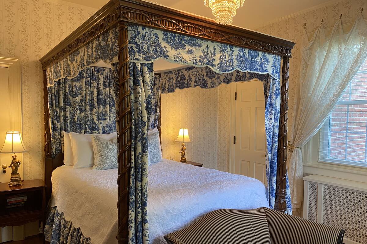 A four-poster bed with blue floral curtains, a wooden headboard, and white bedding. There is a nightstand with a lamp and a statue, and a striped armchair in the foreground.