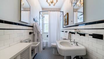 A bathroom with white tiled walls, a black and white patterned floor, a chandelier, a bathtub, a toilet, and a sink.
