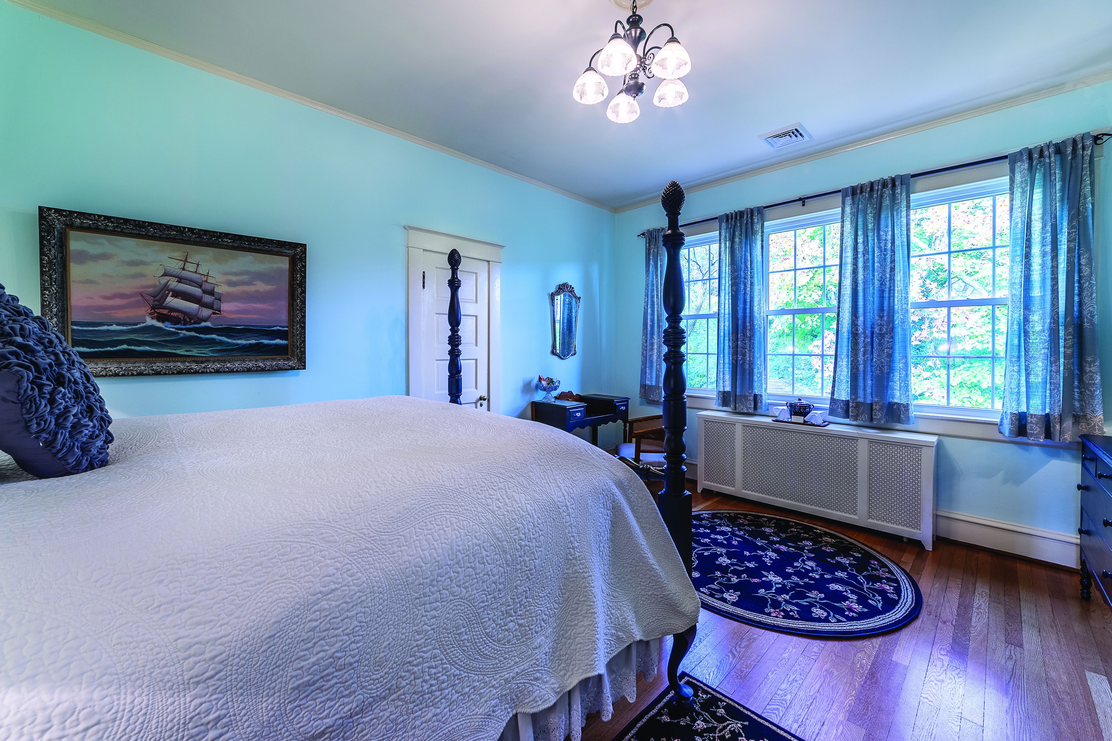 Une chambre à coucher avec un grand lit, un tableau représentant un navire, une fenêtre avec des rideaux et un radiateur.